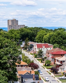 A suburban neighborhood with rows of houses featuring diverse architectural styles, surrounded by lush green trees. In the background, a tall apartment building rises against a backdrop of distant hills and a sky filled with fluffy clouds.