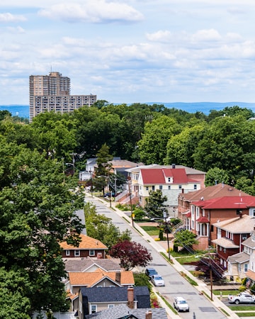 A suburban neighborhood with rows of houses featuring diverse architectural styles, surrounded by lush green trees. In the background, a tall apartment building rises against a backdrop of distant hills and a sky filled with fluffy clouds.