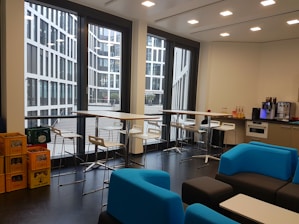 A modern office kitchen area with large windows showcasing a view of a contemporary glass building outside. Inside, there are tall tables with bar stools, a kitchen counter with a coffee machine, several bottles of drinks, and shelves on the right. Blue and black lounge chairs are arranged around low tables. On the left, there are crates filled with beverages.