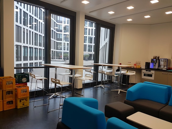 A modern office kitchen area with large windows showcasing a view of a contemporary glass building outside. Inside, there are tall tables with bar stools, a kitchen counter with a coffee machine, several bottles of drinks, and shelves on the right. Blue and black lounge chairs are arranged around low tables. On the left, there are crates filled with beverages.