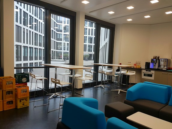 A modern office kitchen area with large windows showcasing a view of a contemporary glass building outside. Inside, there are tall tables with bar stools, a kitchen counter with a coffee machine, several bottles of drinks, and shelves on the right. Blue and black lounge chairs are arranged around low tables. On the left, there are crates filled with beverages.
