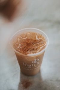 A plastic cup filled with iced coffee topped with a dusting of cinnamon. The cup has a logo and text printed on it and is placed on a light-colored surface.