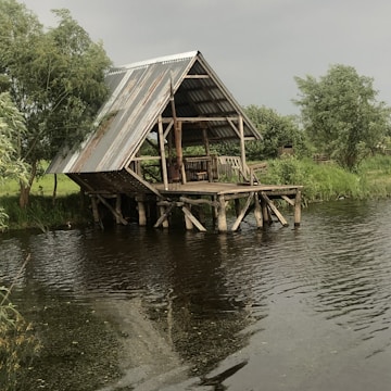 A rustic wooden shelter with a tin roof stands on a small platform supported by wooden stilts overhanging a body of water. The surrounding area is lush with greenery, including trees and grasses. The structure appears weathered but sturdy, with a simple chair and some other small furniture under the shelter.