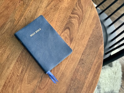 A blue Holy Bible with gold lettering lies on a wooden table. A matching blue ribbon bookmarks part of the book. The table has a rich, brown wood grain texture. In the background, part of a black chair and a textured white rug are visible.