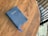 A blue Holy Bible with gold lettering lies on a wooden table. A matching blue ribbon bookmarks part of the book. The table has a rich, brown wood grain texture. In the background, part of a black chair and a textured white rug are visible.