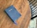 A blue Holy Bible with gold lettering lies on a wooden table. A matching blue ribbon bookmarks part of the book. The table has a rich, brown wood grain texture. In the background, part of a black chair and a textured white rug are visible.
