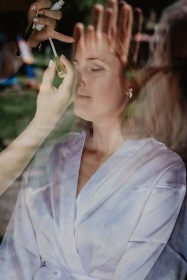 A woman appears to be having her makeup applied, with multiple hands visible holding cosmetic tools. The image is taken through a reflective surface, creating an overlay effect that mixes the reflection with the scene inside.