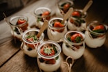 A collection of small glass jars filled with a creamy dessert topped with fresh strawberries and mint leaves. The jars are arranged on a rustic wooden surface, each tied with a piece of twine and includes a small wooden spoon.