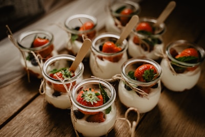 A collection of small glass jars filled with a creamy dessert topped with fresh strawberries and mint leaves. The jars are arranged on a rustic wooden surface, each tied with a piece of twine and includes a small wooden spoon.
