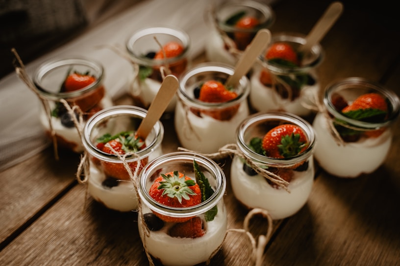 A collection of small glass jars filled with a creamy dessert topped with fresh strawberries and mint leaves. The jars are arranged on a rustic wooden surface, each tied with a piece of twine and includes a small wooden spoon.