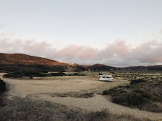 A solitary RV is parked in a vast, open landscape with rolling hills in the background. The ground is covered in dry grass and patches of low shrubs. The sky is filled with soft, diffused clouds as the sun sets, casting a warm hue over the scene.