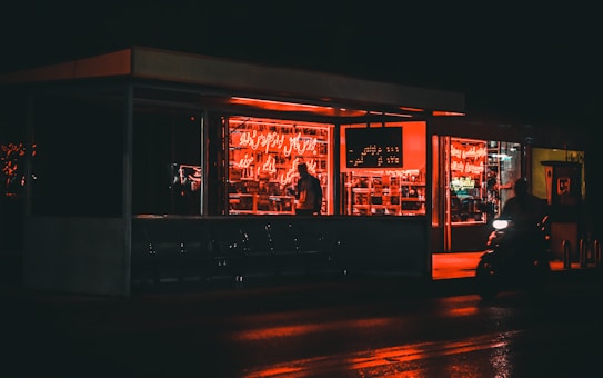 A dimly lit diner or cafe with red neon lights illuminating the interior. The setting appears to be at night with reflections of lights on the wet pavement outside. There is a person sitting inside and another riding a motorcycle past the establishment.