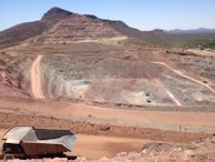 A large open-pit mine is set against a backdrop of arid hills and a clear blue sky. The terraced excavation has visible layers of reddish-brown soil and gray rock. Several large mining trucks navigate the wide dirt roads within the pit.