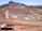 A large open-pit mine is set against a backdrop of arid hills and a clear blue sky. The terraced excavation has visible layers of reddish-brown soil and gray rock. Several large mining trucks navigate the wide dirt roads within the pit.