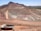 A large open-pit mine is set against a backdrop of arid hills and a clear blue sky. The terraced excavation has visible layers of reddish-brown soil and gray rock. Several large mining trucks navigate the wide dirt roads within the pit.
