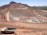 A large open-pit mine is set against a backdrop of arid hills and a clear blue sky. The terraced excavation has visible layers of reddish-brown soil and gray rock. Several large mining trucks navigate the wide dirt roads within the pit.