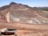 A large open-pit mine is set against a backdrop of arid hills and a clear blue sky. The terraced excavation has visible layers of reddish-brown soil and gray rock. Several large mining trucks navigate the wide dirt roads within the pit.
