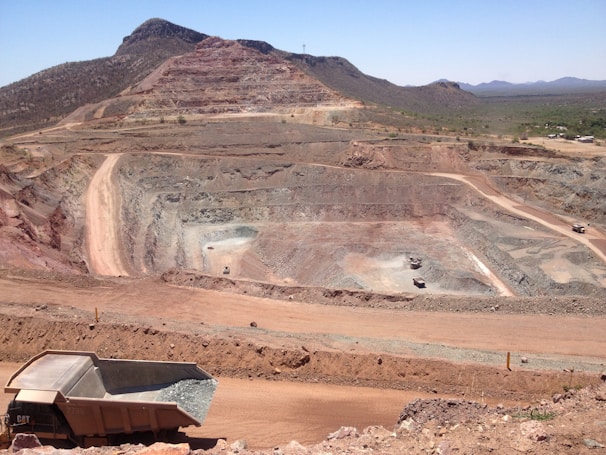 A large open-pit mine is set against a backdrop of arid hills and a clear blue sky. The terraced excavation has visible layers of reddish-brown soil and gray rock. Several large mining trucks navigate the wide dirt roads within the pit.