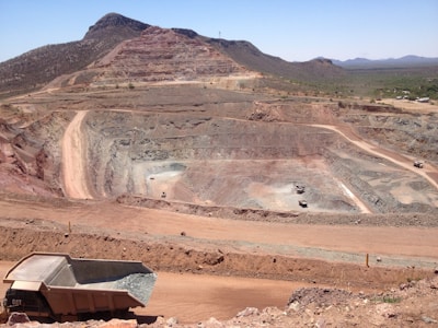 A large open-pit mine is set against a backdrop of arid hills and a clear blue sky. The terraced excavation has visible layers of reddish-brown soil and gray rock. Several large mining trucks navigate the wide dirt roads within the pit.