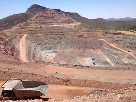 A large open-pit mine is set against a backdrop of arid hills and a clear blue sky. The terraced excavation has visible layers of reddish-brown soil and gray rock. Several large mining trucks navigate the wide dirt roads within the pit.