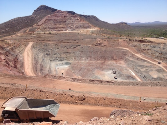 A large open-pit mine is set against a backdrop of arid hills and a clear blue sky. The terraced excavation has visible layers of reddish-brown soil and gray rock. Several large mining trucks navigate the wide dirt roads within the pit.