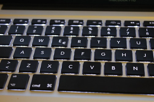 A close-up view of a computer keyboard featuring black keys with white letters and symbols. The keys are backlit, creating a soft glow around each one. The keyboard layout includes a row of function keys at the top, along with standard QWERTY keys.