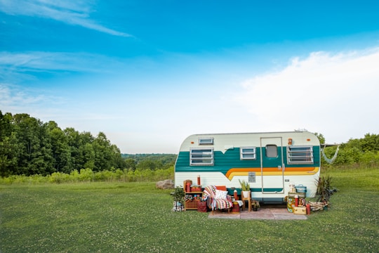 A vintage camper trailer is parked on a grassy field with a backdrop of lush green trees and a bright blue sky. The trailer is decorated with colorful stripes and surrounded by various outdoor items such as plants, a patterned blanket, and stacked crates, creating a cozy and rustic setting.