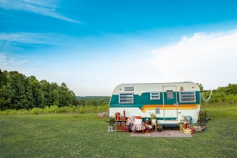A vintage camper trailer is parked on a grassy field with a backdrop of lush green trees and a bright blue sky. The trailer is decorated with colorful stripes and surrounded by various outdoor items such as plants, a patterned blanket, and stacked crates, creating a cozy and rustic setting.