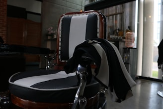 A vintage-style barber chair with black and white striped upholstery sits in a dimly lit room. A striped cloth is draped over one arm of the chair. The background features a concrete wall, a shelf with decorative items, and a large water dispenser. The room has industrial decor with metal and wood elements.