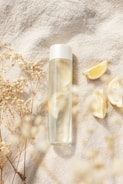 A transparent water bottle with a white cap placed on a textured fabric surface, surrounded by dried flowers and lemon slices, creating a calm and refreshing scene.