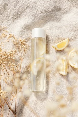 A transparent water bottle with a white cap placed on a textured fabric surface, surrounded by dried flowers and lemon slices, creating a calm and refreshing scene.