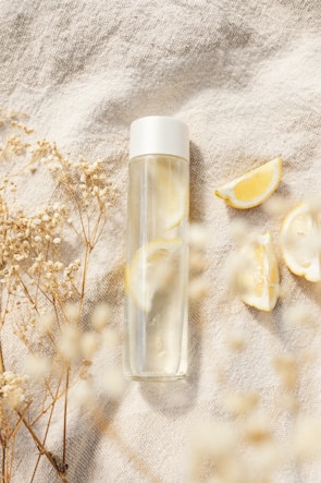 A transparent water bottle with a white cap placed on a textured fabric surface, surrounded by dried flowers and lemon slices, creating a calm and refreshing scene.
