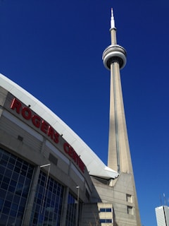 A tall communications and observation tower stands prominently against a clear blue sky. In the foreground, part of a large stadium roof is visible with large red letters spelling 'ROGERS CENTRE'.