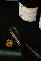 Two wine bottles with elegant labels, the label showcasing a minimalist design and the number '20' prominently visible. A small yellow flower lies next to one of the bottles on a dark surface.