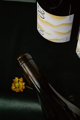 Two wine bottles with elegant labels, the label showcasing a minimalist design and the number '20' prominently visible. A small yellow flower lies next to one of the bottles on a dark surface.