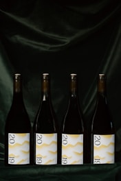 Four wine bottles are evenly lined up against a dark, velvety background. Each bottle features a label displaying the year 2019 with a white background and a yellow zigzag pattern.