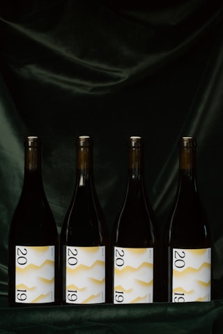 Four wine bottles are evenly lined up against a dark, velvety background. Each bottle features a label displaying the year 2019 with a white background and a yellow zigzag pattern.