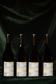 Four wine bottles are evenly lined up against a dark, velvety background. Each bottle features a label displaying the year 2019 with a white background and a yellow zigzag pattern.