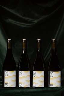Four wine bottles are evenly lined up against a dark, velvety background. Each bottle features a label displaying the year 2019 with a white background and a yellow zigzag pattern.