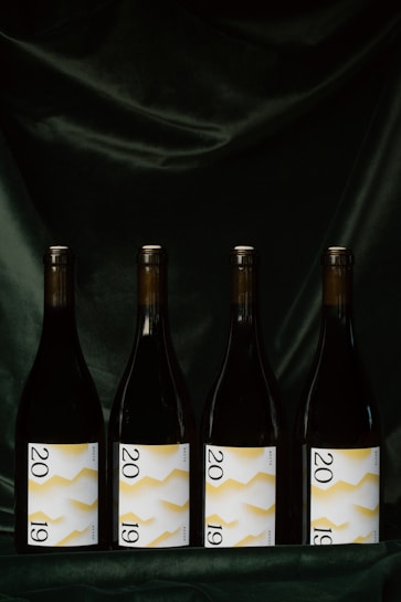 Four wine bottles are evenly lined up against a dark, velvety background. Each bottle features a label displaying the year 2019 with a white background and a yellow zigzag pattern.