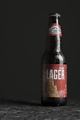 A bottle of Granville Island Brewing lager stands upright on a textured surface. The bottle is dark with condensation droplets on its exterior, suggesting it is cold. The label is predominantly red with white text that reads 'Island Lager'.