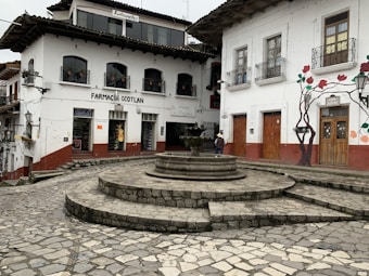 A quaint town square with stone-paved streets and a central stone fountain. Traditional white buildings with red accents surround the square, featuring ornate iron window railings and wood-framed doors. A mural with floral designs adorns one wall, and a few people are walking on the street, giving the scene a quiet, rustic charm.