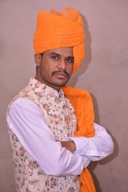 A man stands with arms crossed, wearing a traditional orange turban and a beige floral-patterned waistcoat over a white shirt. The background is a plain wall.