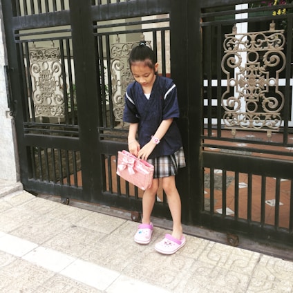 A young girl stands in front of an ornate black metal gate. She is wearing a dark blue top and plaid skirt, holding a pink bag with a bow. She has pink and white slippers and a pink wristwatch. Her hair is tied back, and she appears to be focused on her bag.