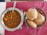 A table with a steel bowl filled with a spicy Indian curry, garnished with herbs and containing chunks of potatoes. Next to it, a basket with several pieces of puffed Indian bread known as puris, on a red cloth.