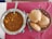A table with a steel bowl filled with a spicy Indian curry, garnished with herbs and containing chunks of potatoes. Next to it, a basket with several pieces of puffed Indian bread known as puris, on a red cloth.