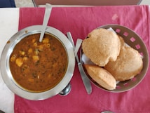 A table with a steel bowl filled with a spicy Indian curry, garnished with herbs and containing chunks of potatoes. Next to it, a basket with several pieces of puffed Indian bread known as puris, on a red cloth.