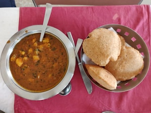 A table with a steel bowl filled with a spicy Indian curry, garnished with herbs and containing chunks of potatoes. Next to it, a basket with several pieces of puffed Indian bread known as puris, on a red cloth.