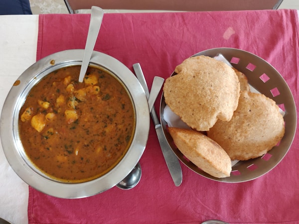 A table with a steel bowl filled with a spicy Indian curry, garnished with herbs and containing chunks of potatoes. Next to it, a basket with several pieces of puffed Indian bread known as puris, on a red cloth.