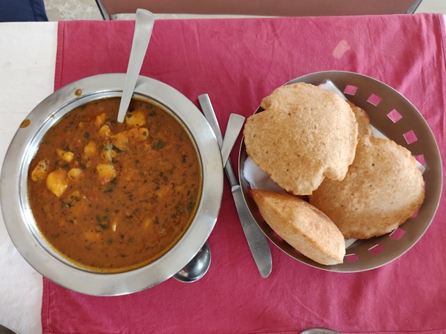 A table with a steel bowl filled with a spicy Indian curry, garnished with herbs and containing chunks of potatoes. Next to it, a basket with several pieces of puffed Indian bread known as puris, on a red cloth.